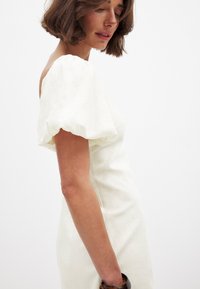 Weißes Off-Shoulder-Kleid mit Puffärmeln, aus leichtem Stoff gefertigt. Enganliegendes Oberteil mit einer glatten, leicht strukturierten Oberfläche. Inklusive eines Armreifs.