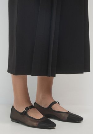 Personne portant des ballerines noires transparentes de type Mary Jane avec des sangles à boucle et une jupe midi noire, se tenant sur un sol blanc devant un fond uni.