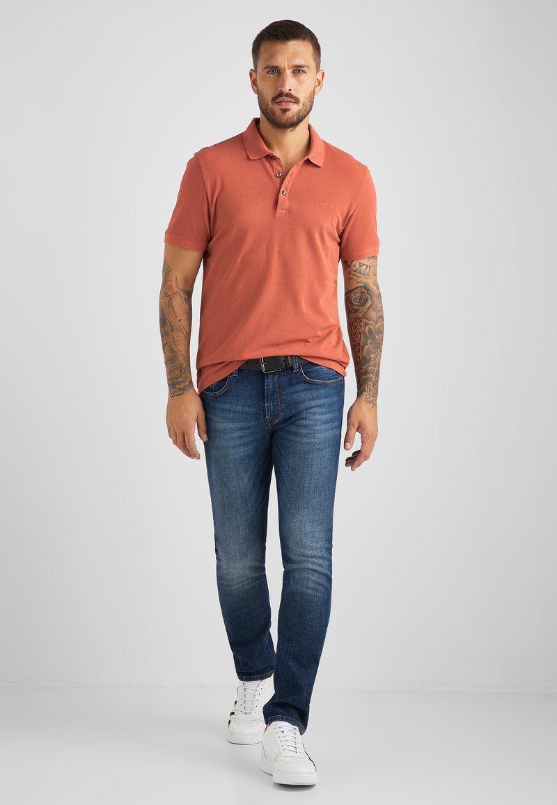 Pánská oranžová polo košile s krátkým rukávem a třemi knoflíky, sladěná s tmavě modrými slim-fit džíny a bílými teniskami. Na pažích jsou viditelné tetování.