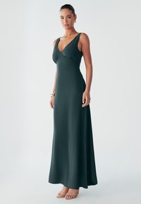 Naine, kandes varrukateta tumerohelist põrandani ulatuvat kleiti ja beeže kontsakingi, seisab ühevärvilise heleda tausta ees.