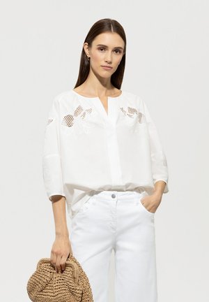 Weiße Baumwollbluse mit gestickten Blumendetails und einem Ausschnitt mit Schlitz. Sie hat Dreiviertelärmel und eine lockere Passform. Kombiniert mit weißen Hosen.