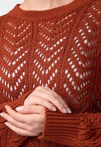 Gebreide trui in verbrand oranje, met een chevronpatroon. Geverfde open structuur met een geribde kraag en manchetten. Handen voor zich gevouwen.