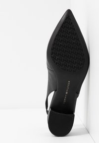 Semelle de chaussure à talon noir à bout pointu avec motif texturé et logo doré Tommy Hilfiger sur fond blanc.
