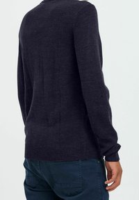 Mann in einem dunkelblauen Pullover und blauen Hosen, von hinten gezeigt, mit der rechten Hand entspannt an der Seite.