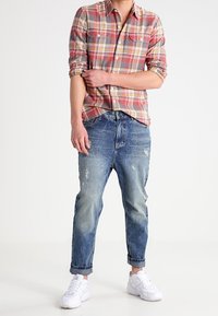 Camicetta a quadri rossi e grigi con bottoni, jeans blu délavé e sneakers bianche. Vestibilità rilassata con maniche arrotolate sui jeans e design sovrapposto.