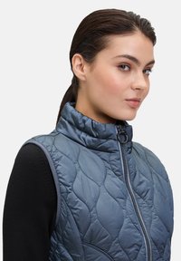 Gevoerde, mouwloze vest in donkerblauw met een hoge kraag, gewatteerde textuur, zijrits en grijze accenten langs de armsgaten.
