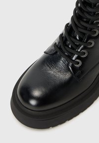 Gros plan d'une botte en cuir noir avec des lacets épais et des œillets, avec une finition brillante et une semelle épaisse sur fond blanc.