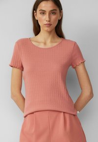 Rosa geripptes T-Shirt mit kurzem Ärmel und Rundhalsausschnitt, das ein strukturiertes Muster und eine gewellte Kante an den Ärmeln aufweist. Weicher Stoff, figurbetontes Design.