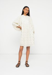 Vestido branco com um padrão texturizado em zigzag, mangas longas e volumosas, um decote redondo e um corte descontraído. Usado com sandálias pretas de tiras.