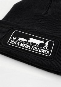 Black knit beanie featuring a white rectangular patch with illustrations of a chicken, pig, cow, and a walking figure, alongside the text "ICH & MEINE FOLGENDER."