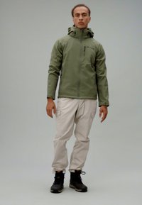 Oliwkowa kurtka z wysokim kołnierzem i zamek błyskawiczny, w zestawieniu z jasnobeżowymi spodniami cargo i czarnymi butami, stojąca na neutralnym tle.