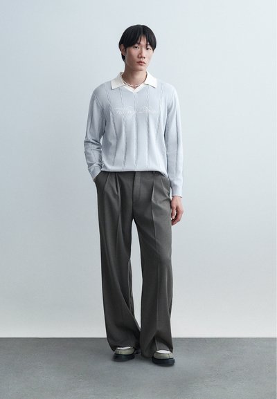 Ασιάτης άνδρας στέκεται μπροστά σε απλό τοίχο, φορώντας ανοιχτό μπλε πλεκτό πουλόβερ με λευκό γιακά, γκρι παντελόνι με φαρδιά πόδια, μαργαριταρένιο κολιέ και αθλητικά παπούτσια.