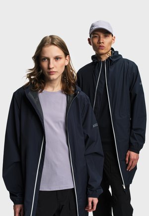 Jeune femme et homme portant des vestes noires zippées ; la femme porte un t-shirt lavande, l'homme une casquette grise et un pantalon noir, fond neutre.