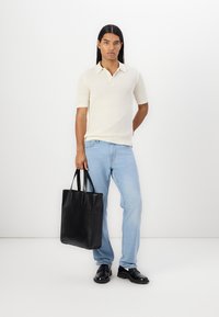 Svart lær tote med minimalistisk design, to solide håndtak og en glatt tekstur. Modell iført en kremfarget ribbestrikket polo og lyseblå jeans.