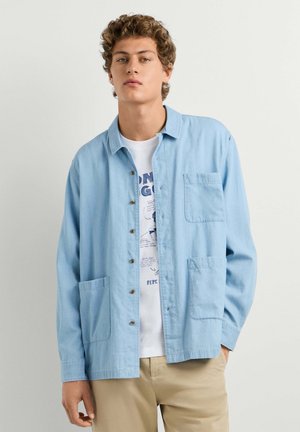 Joven con pelo rizado que lleva una camisa azul claro abotonada sobre una camiseta blanca con estampado y pantalones beige, de pie frente a un fondo liso.