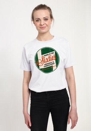 Kvinde med blondt hår iført en hvid T-shirt med teksten "Født i tresserne, original og urestaureret" med et grønt og beige cirkeldesign.