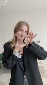 Gullnøkkelring med en anheng og en svart vedheng, holdt av en person som har på seg en svart blazer og lysegrå skjorte, mot en nøytral bakgrunn.