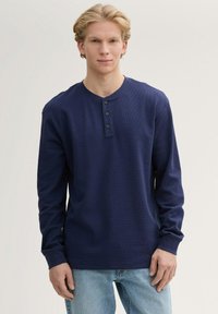 Navyblaues Langarm-Henley-Shirt aus strukturiertem Stoff mit einer Knopfleiste mit drei Knöpfen und gerippten Bündchen. Getragen mit hellblauen Jeans.
