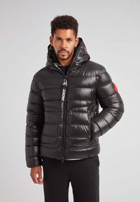 Giubbotto piumino nero con cappuccio aderente, design trapuntato, texture lucida, chiusura frontale con zip, tasche laterali e un accento rosso sulla manica sinistra.