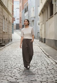 Krémový top s krátkým rukávem a volné hnědé kalhoty na šňůrku. Modelka prochází po dlážděné ulici mezi budovami.