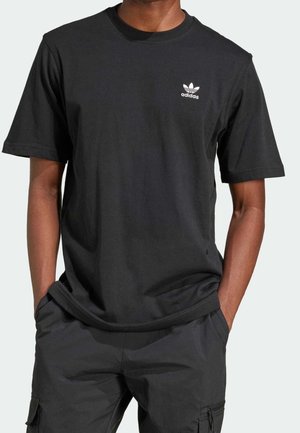 Mand iført en sort Adidas t-shirt med lille hvidt logo på brystet, kombineret med sorte cargobukser, hænderne i lommerne.