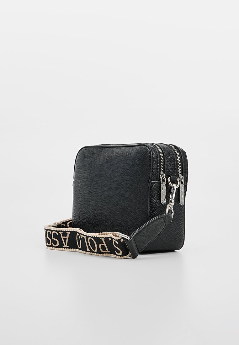 Bolso cruzado de cuero negro con dos compartimentos con cremallera y una correa estampada que presenta "POLO ASSN" en color crema sobre negro.