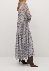 Nainen, jolla on päällään pitkä, kuviollinen, läpikuultava mekko, jossa on pitkät hihat, ja ruskeat teräväkärkiset korkokengät, seisoo tasaista taustaa vasten.