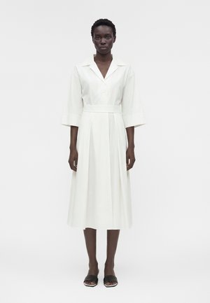 Žena v bielych midi šatách s 3/4 rukávmi, golierom, širokým pásom a čiernymi lodičkami s otvorenou špičkou, stojaca na jednoduchom pozadí.