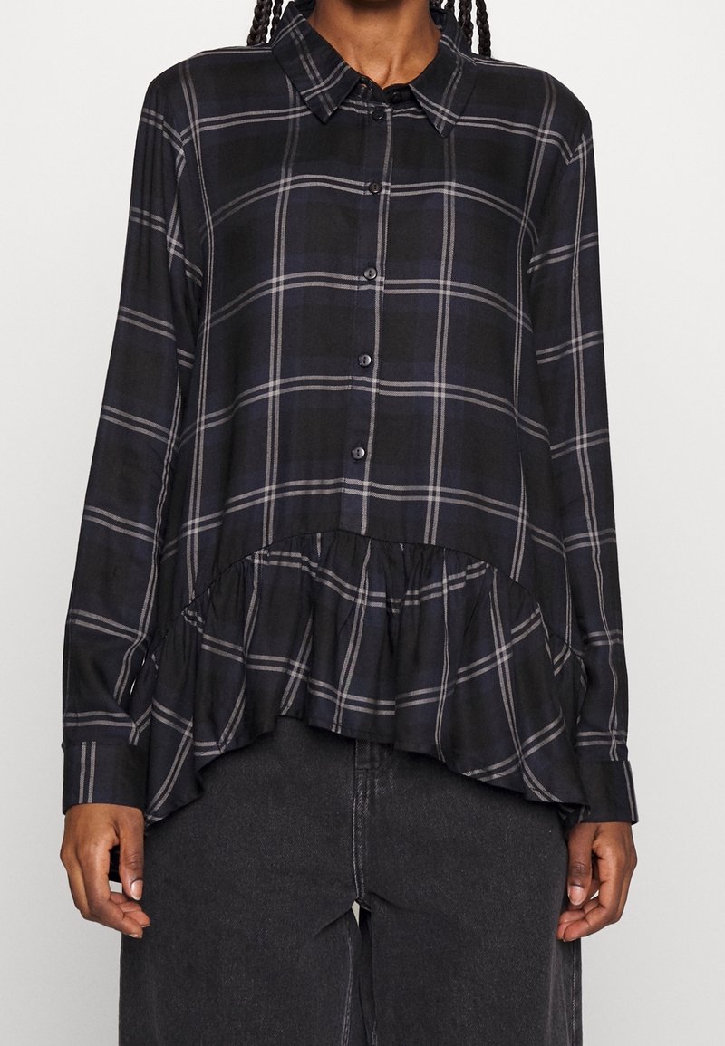 Chemise à manches longues à carreaux noirs avec boutons sur le devant, ourlet à volants et col ; présente des rayures blanches et grises dans un motif à carreaux.