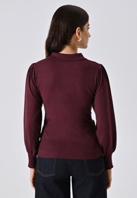 Donna vista da dietro che indossa un maglione bordeaux a maniche lunghe con colletto e jeans blu scuro, su uno sfondo grigio uniforme.
