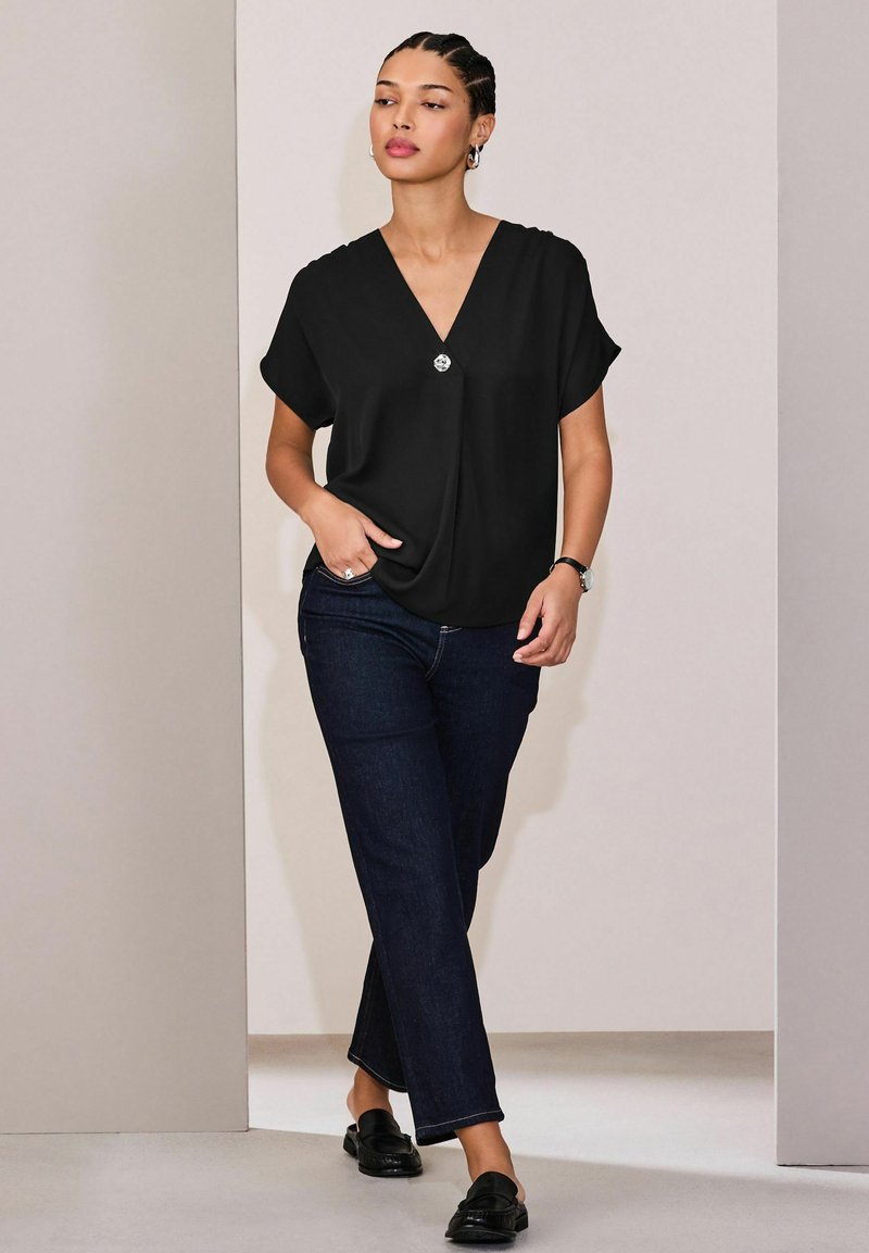 Črna bluza z V-izrezom in kratkimi rokavi, s srebrnim gumbom na prsih. Oblečena z temnimi kavbojkami in črnimi natikači.