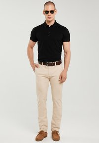 Mann trägt schwarzes Poloshirt, beige Hose, braunen Gürtel, braune Schuhe und Sonnenbrille, steht vor hellem Hintergrund.