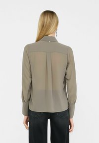 Dlouhá, button-down košile v světle olivové barvě, s průhlednou zadní částí, zaobleným dolním lemem a nařasenými manžetami. Jednoduchý detail límce.
