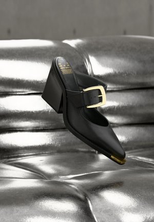 Mule en cuir noir avec bout pointu, boucle dorée et talon bloc suspendue au-dessus d'une surface rembourrée argentée brillante.