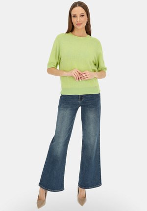 Kobieta stoi, ubrana w jasnziezielony sweter z krótkim rękawem, szerokie niebieskie jeansy i beżowe szpiczaste szpilki, na tle jednolitego tła.