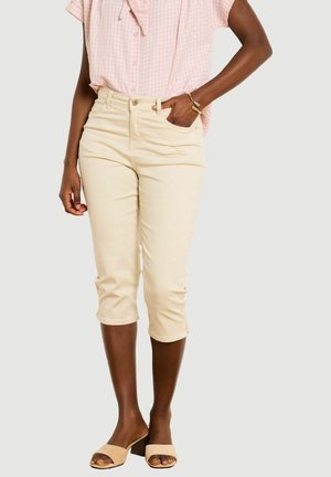 Kobieta ubrana w beżowe krótkie spodnie, różową bluzkę z krótkim rękawem i beżowe sandały na szerokim obcasie z otwartymi palcami, ręka spoczywa w kieszeni.