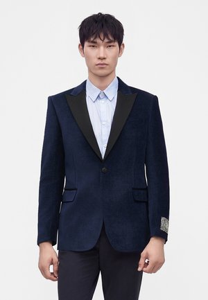 Jeune homme aux cheveux foncés porte un blazer bleu marine à revers noirs, une chemise bleu clair à col et un pantalon foncé sur fond uni.