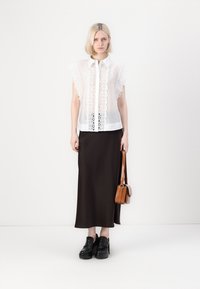 Blusa bianca trasparente con dettagli in pizzo, abbinata a una gonna maxi marrone. La modella indossa scarpe platform nere e tiene in mano una piccola borsa marrone.