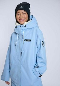 Lyseblå isoleret vinterjakke med hætte, der har en hvid fleeceforing, justerbare snore, sorte detaljer og mærkning.