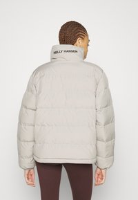 Vyrážková, světle šedá bunda s vysokým límcem a černým logem "Helly Hansen" na zadní straně. Obsahuje vodorovné prošívané části a elastické manžety.
