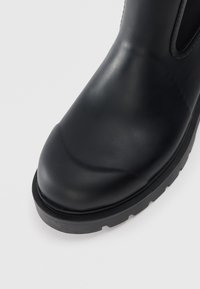 Botte en caoutchouc noir à cheville avec un bout arrondi, finition mate et semelle texturée, dotée de panneaux latéraux élastiques pour un enfilage facile.