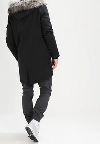 Svart parkas med huva i fuskpäls, lång klippning och strömlinjeformat utseende, ihop med grå cargobyxor och vita sneakers.