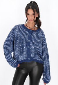 Modrý pletený cardigan s volným střihem, kulatým výstřihem, sedmi velkými knoflíky a texturovaným designem, kombinovaný s černými legínami.