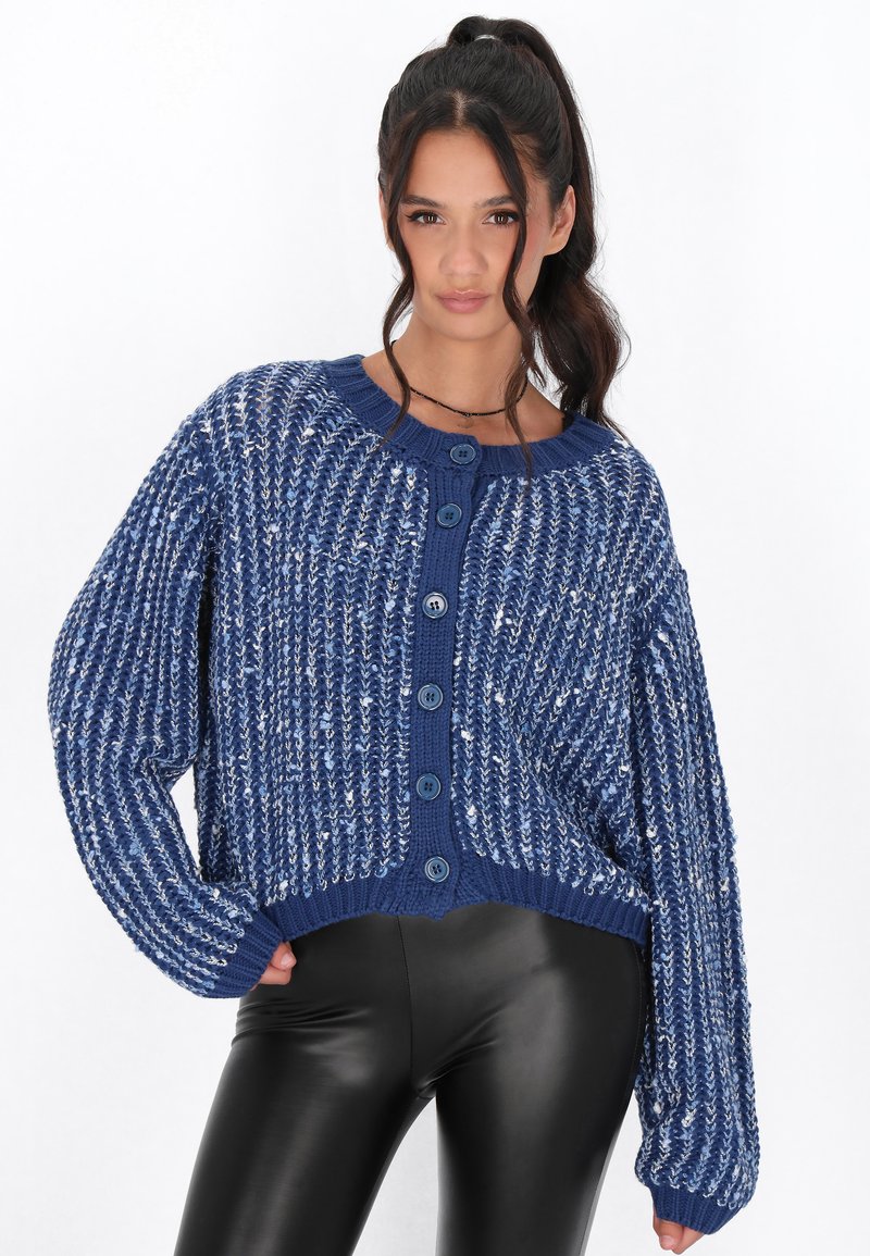 Modrý pletený cardigan s volným střihem, kulatým výstřihem, sedmi velkými knoflíky a texturovaným designem, kombinovaný s černými legínami.