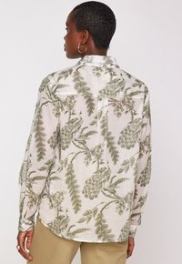Camicia a maniche lunghe in tessuto leggero con stampa botanica che presenta motivi di pavoni verdi su uno sfondo crema. Collo classico e polsini con bottoni.