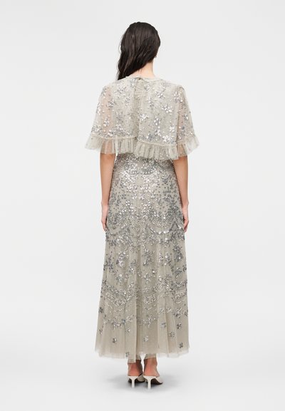 Γυναίκα που φοράει μακριά διάφανη φορεσιά με ασημένιες λουλουδένιες και παγιετέ λεπτομέρειες, στέκεται με την πλάτη σε απλό φόντο.
