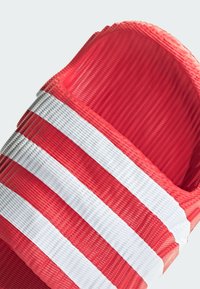 Close-up of a red slide sandal with three white horizontal stripes on the wide strap against a light background.