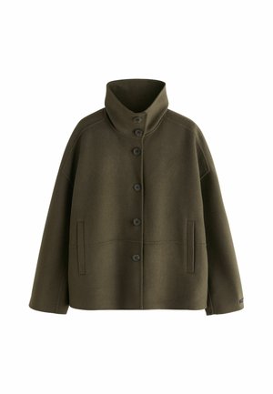 Frau von hinten gesehen, trägt eine locker sitzende dunkelgrüne Jacke mit hohem Kragen bei weichem Innenlicht.