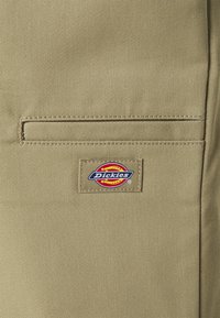 Brūns audums ar redzamu plāksteri, kurā redzams uzraksts "Dickies" zili, sarkani un dzelteni. Iekļauj horizontālu kabatu virs plākstera.