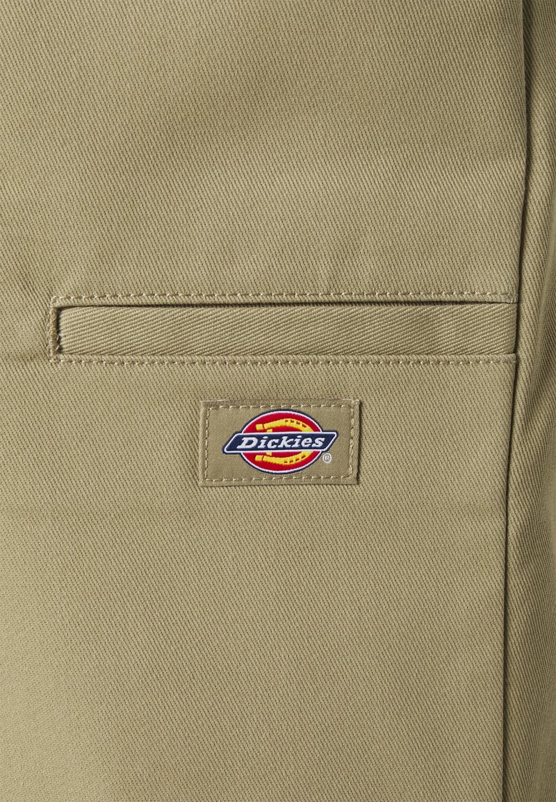 Tan fabric with a visible patch label reading "Dickies" in blue, red, and yellow. Features a horizontal welt pocket above the label.